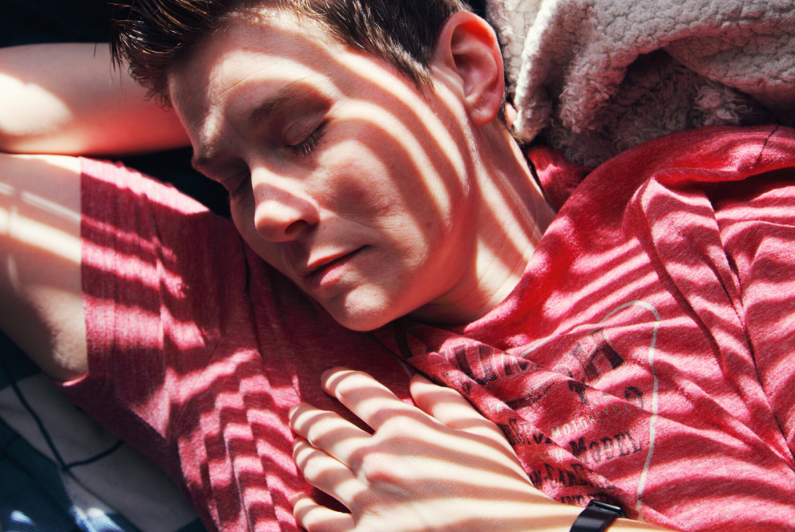 Kuvassa lähikuva henkilöstä, punainen paita. Henkilö nukkuu ja aurinko paistaa sälekaihtimien tms. välistä