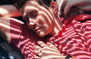 Kuvassa lähikuva henkilöstä, punainen paita. Henkilö nukkuu ja aurinko paistaa sälekaihtimien tms. välistä