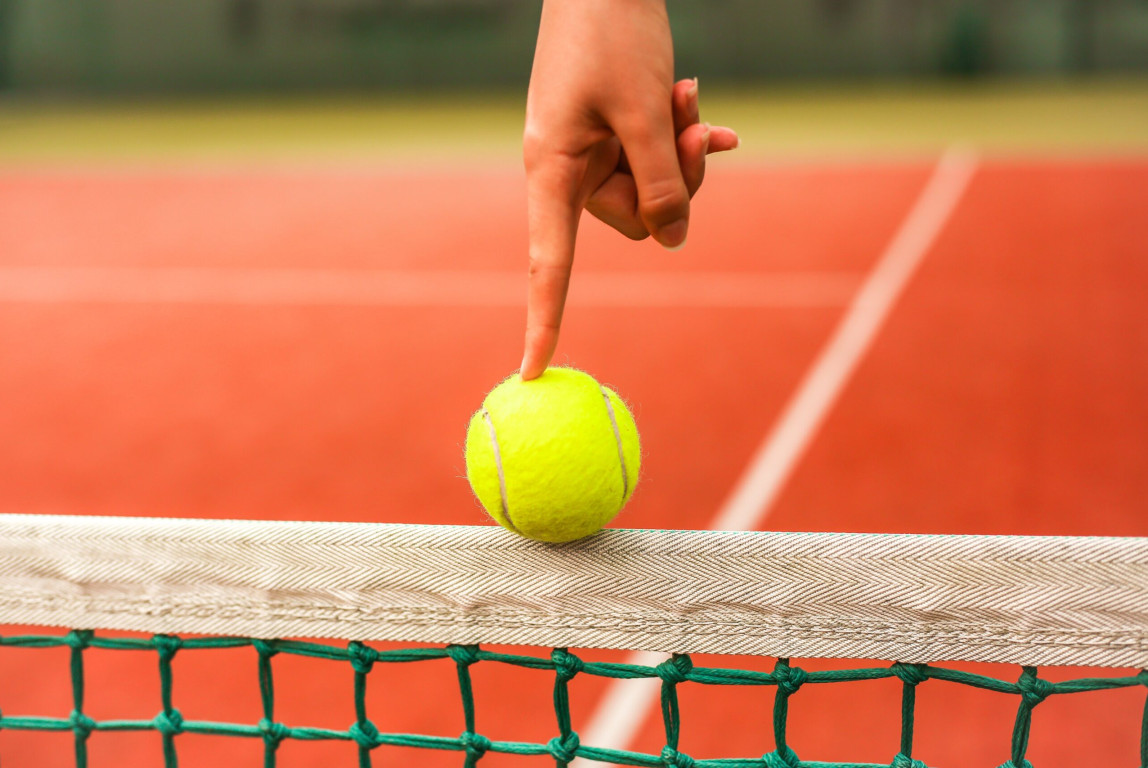 Kuvassa tennispallo, jota ihmisen sormi tasapainottelee verkon päällä.