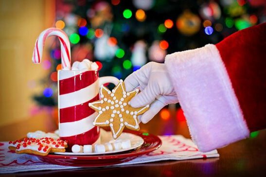 Joulupukin käsin ottaa lautaselta tähden muotoisen piparin. Lautasella on pipareita, vaahtokarkkeja ja punavalkoinen muki, jossa karkkikeppi pystyssä.