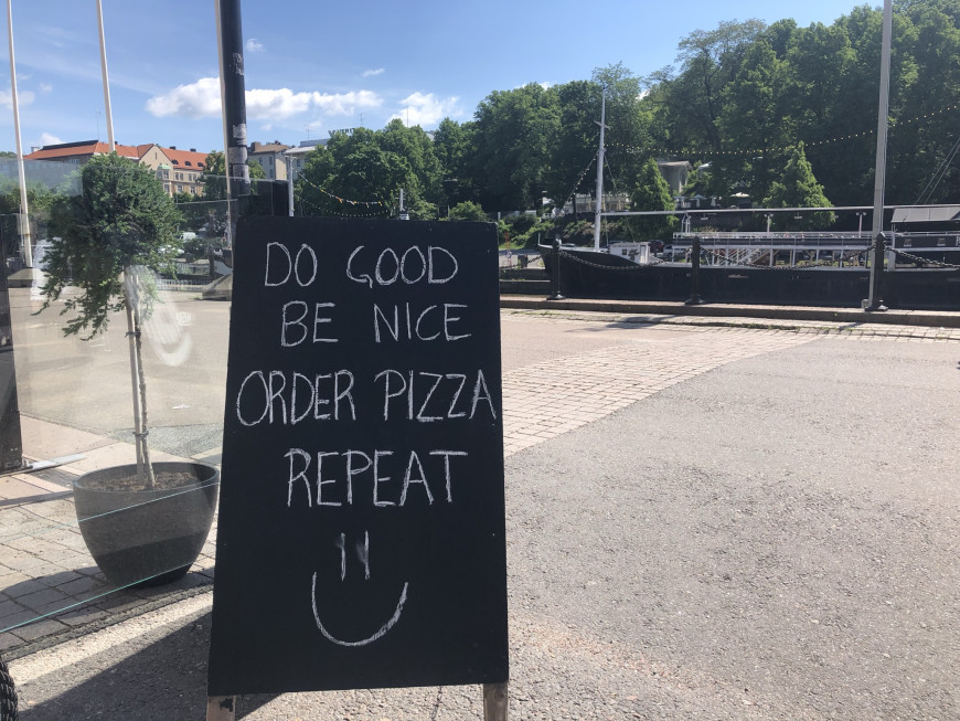 Kuvassa kesäisessä maisemassa musta mainoskyltti, jossa lukee valkoisilla kirjaimilla: do good, be nice, order pizza, repeat.