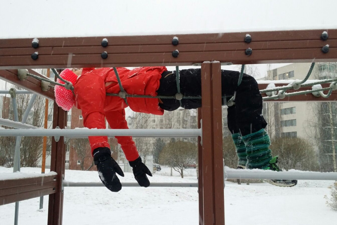 Kuvassa ihminen makaa selällään raajat roikkuen lasten kiipeilytelineessä lumisateessa.